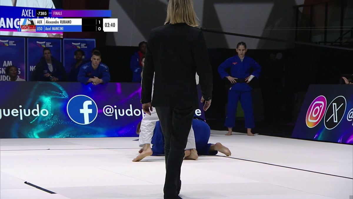 Plan large d'un combat sur tatami blanc, bandeau de score en haut à gauche.